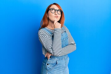 Beautiful redhead woman wearing casual clothes and glasses thinking concentrated about doubt with...