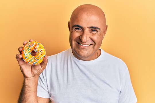 Mature middle east man holding tasty colorful doughnut looking positive and happy standing and smiling with a confident smile showing teeth