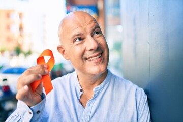 Middle age bald man smiling happy holding orange ribbon leaning on the wall at the city