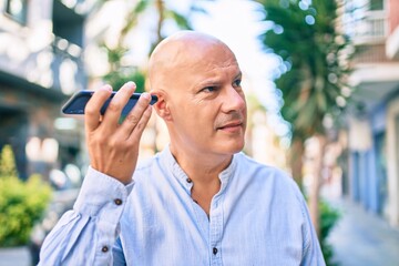 Middle age bald man smiling happy listening audio message using smartphone at the city.