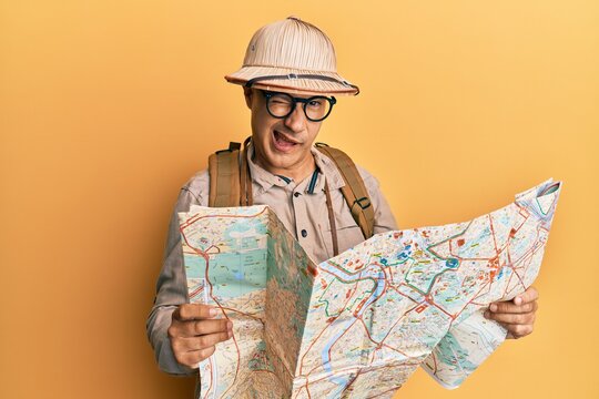 Middle age bald man wearing explorer hat holding map winking looking at the camera with sexy expression, cheerful and happy face.