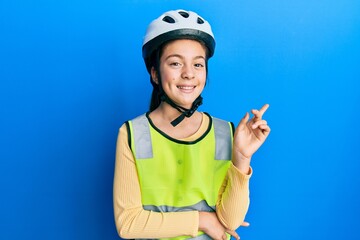 Beautiful brunette little girl wearing bike helmet and reflective vest with a big smile on face, pointing with hand and finger to the side looking at the camera.