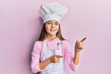 Beautiful brunette little girl wearing professional cook apron and hat smiling and looking at the camera pointing with two hands and fingers to the side.