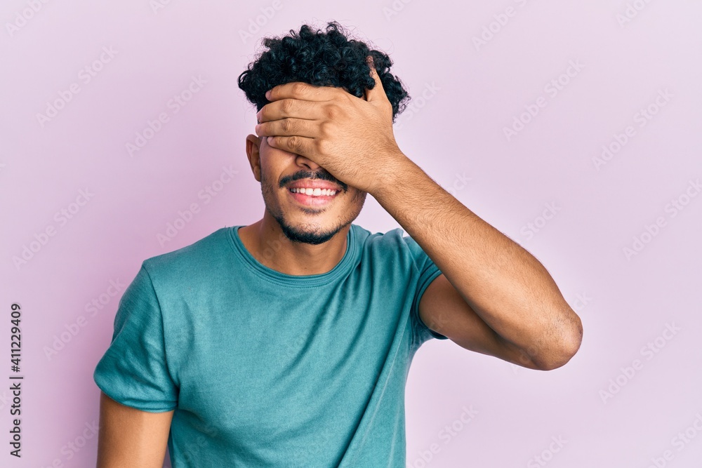 Wall mural Young arab handsome man wearing casual clothes smiling and laughing with hand on face covering eyes for surprise. blind concept.