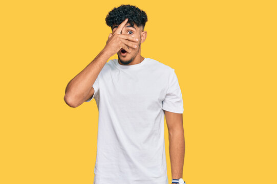 Young arab man wearing casual white t shirt peeking in shock covering face and eyes with hand, looking through fingers with embarrassed expression.