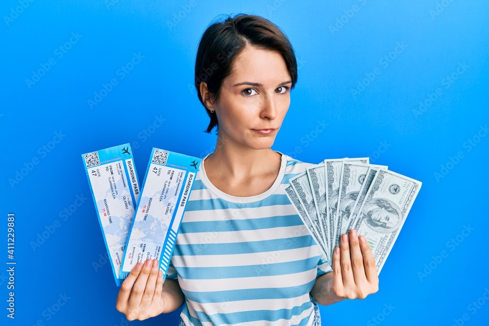 Wall mural Young brunette woman with short hair holding boarding pass and dollars clueless and confused expression. doubt concept. - Wall murals