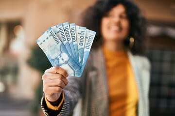 Young hispanic businesswoman smiling happy holding chilean pesos banknotes at the city.