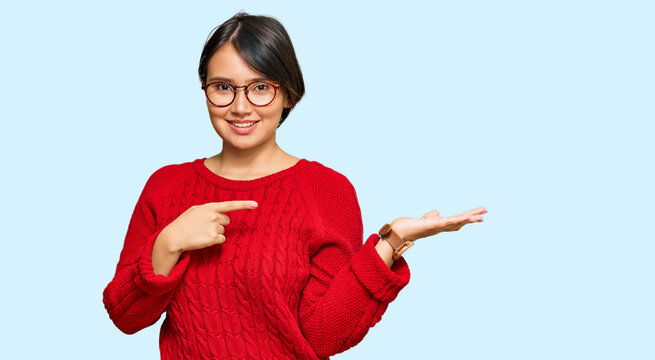 Young beautiful hispanic woman with short hair wearing casual sweater and glasses amazed and smiling to the camera while presenting with hand and pointing with finger.