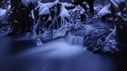 beautifully flowing water with a cascade, tree trunks, frozen branches, snow and icicles. long exposure. blue tint