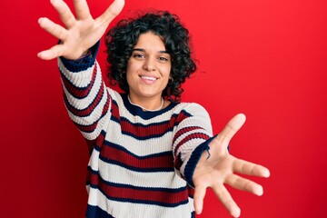 Young hispanic woman with curly hair wearing casual winter sweater looking at the camera smiling with open arms for hug. cheerful expression embracing happiness.