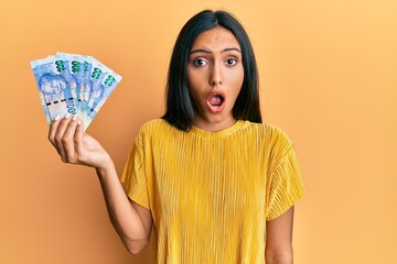 Young brunette woman holding south african 100 rand banknotes scared and amazed with open mouth for surprise, disbelief face