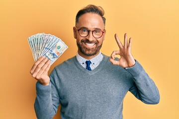 Handsome middle age man holding dollars doing ok sign with fingers, smiling friendly gesturing excellent symbol