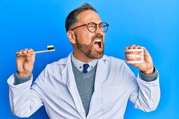 Handsome middle age dentist man holding denture and toothbrush angry and mad screaming frustrated...