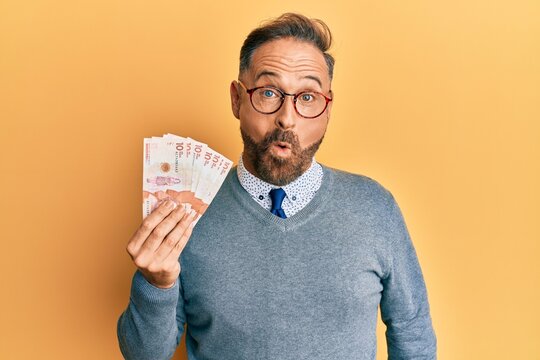 Handsome middle age man holding 10 colombian pesos banknotes scared and amazed with open mouth for surprise, disbelief face