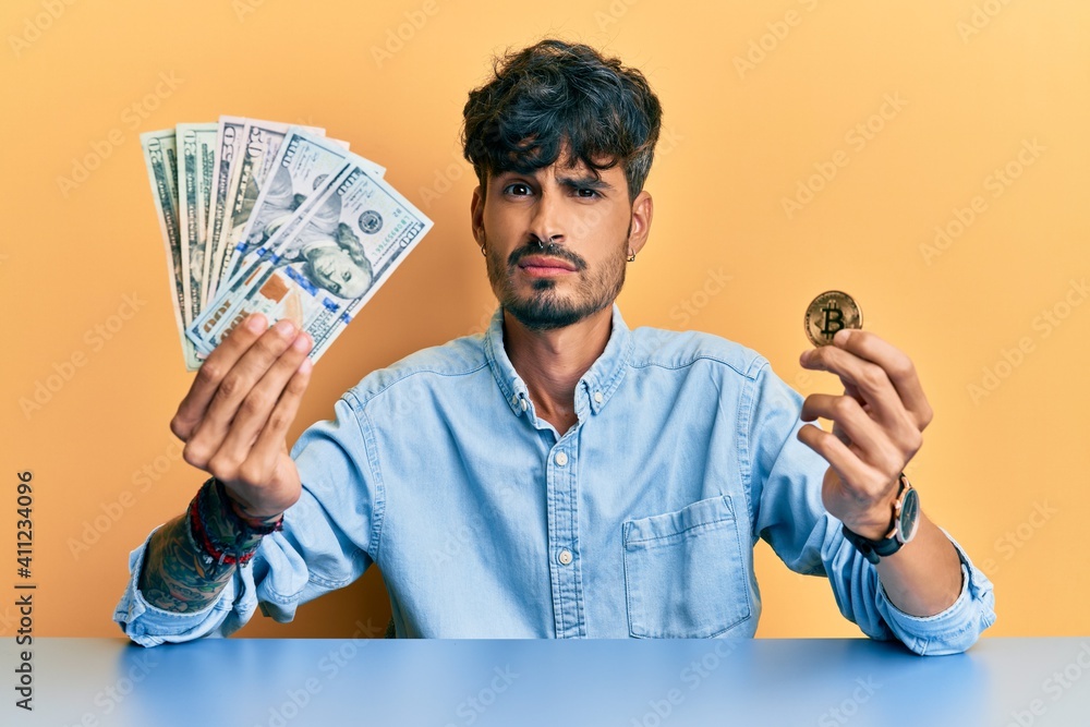 Wall mural Young hispanic man holding dollars and bitcoin clueless and confused expression. doubt concept. - Wall murals