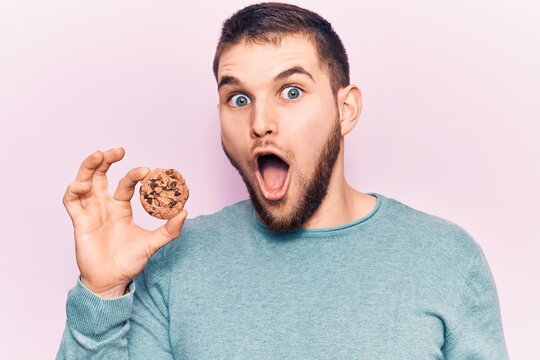 Young handsome man holding cookie scared and amazed with open mouth for surprise, disbelief face