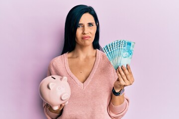 Young caucasian woman holding brazil real banknotes and piggy bank skeptic and nervous, frowning upset because of problem. negative person.