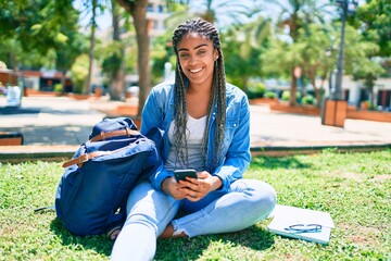 Young african american student woman smiling happy using smartphone sitting on the grass at the...