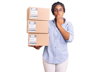 Young african american woman with braids holding delivery packages covering mouth with hand, shocked and afraid for mistake. surprised expression