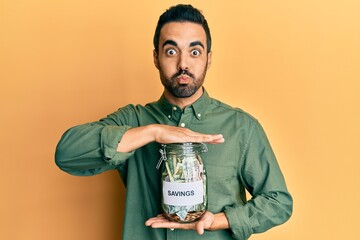 Young hispanic man holding jar with savings puffing cheeks with funny face. mouth inflated with air, catching air.