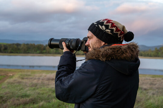 Photo of a attractive and young photographer in the middle of the wild looking through his camera zoom lens. Wearing winter clothes. Explorer, looking for animals