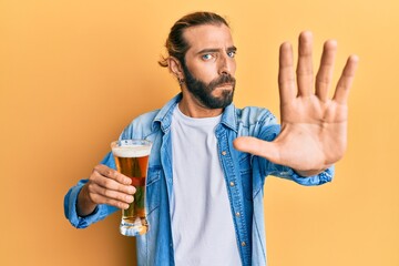 Attractive man with long hair and beard drinking a pint of beer with open hand doing stop sign with serious and confident expression, defense gesture