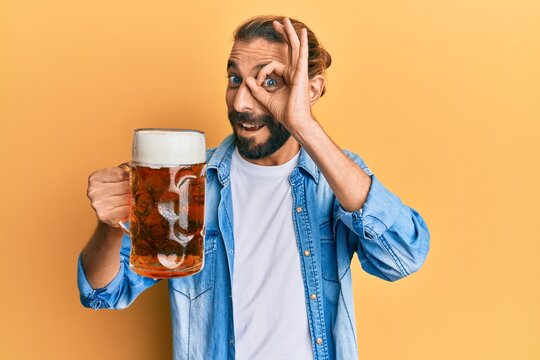 Attractive man with long hair and beard drinking a pint of beer smiling happy doing ok sign with hand on eye looking through fingers