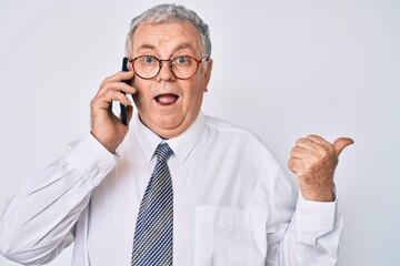 Senior grey-haired man wearing business clothes having conversation talking on the smartphone pointing thumb up to the side smiling happy with open mouth