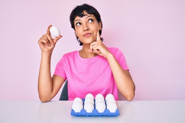 Beautiful brunettte woman showing fresh white eggs serious face thinking about question with hand...