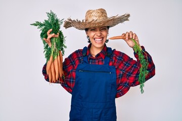 Beautiful brunettte woman wearing farmer clothes holding fresh carrots smiling and laughing hard out loud because funny crazy joke.