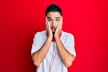 Young man with beard listening to music using headphones afraid and shocked, surprise and amazed expression with hands on face