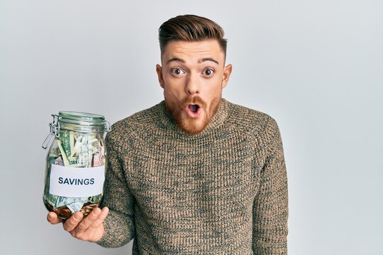 Young redhead man holding jar with savings scared and amazed with open mouth for surprise, disbelief face