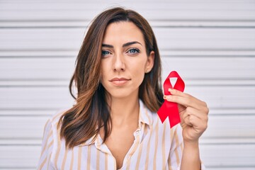 Young hispanic woman with serious expression holding red hiv ribbon standing at the city.