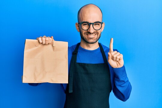 Young hispanic man wearing waiter uniform holding take away paper bag smiling with an idea or question pointing finger with happy face, number one