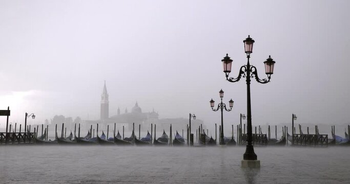 Solemn shot of an empty promenade with boats rocking on stormy water while rain is sweeping across the street. A hazy mist covers a large tower in the background.