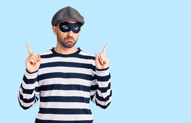 Young handsome man wearing burglar mask pointing up looking sad and upset, indicating direction with fingers, unhappy and depressed.