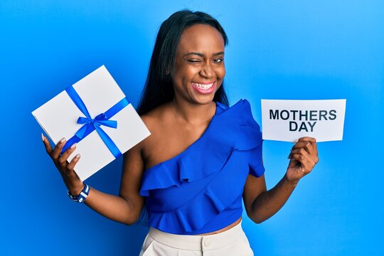 Young African Woman Holding Gift And Mothers Day Text Winking Looking At The Camera With Sexy Expression, Cheerful And Happy Face.