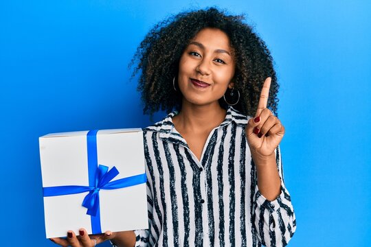 Beautiful african american woman with afro hair holding gift smiling with an idea or question pointing finger with happy face, number one