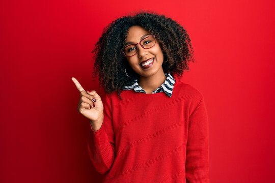Beautiful african american woman with afro hair wearing sweater and glasses with a big smile on face, pointing with hand finger to the side looking at the camera.