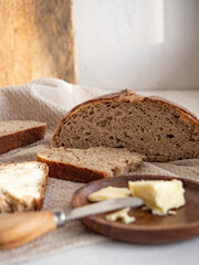 Homemade fresh wheat-rye bread on sourdough on a natural color linen napkin with butter in a wooden plate with a butter knife on a white background in the morning light