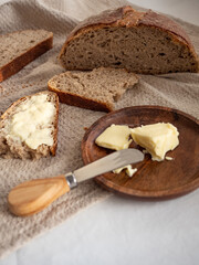 Homemade fresh wheat-rye bread on sourdough on a natural color linen napkin with butter in a wooden plate with a butter knife on a white background in the morning light