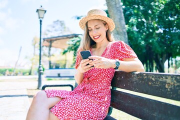 Young blonde woman on vacation using smartphone sitting on bench at the park
