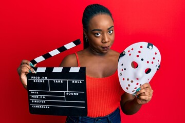 Young african american woman holding video film clapboard and fear mask skeptic and nervous,...