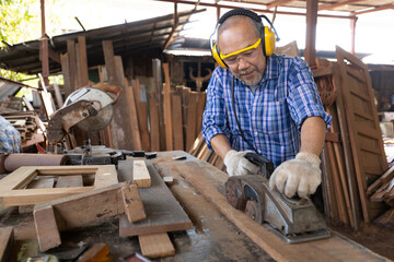 senior asian man carpenter wearing glasses and headphone, using electric wood planers on a piece of wood in workshop - obrazy, fototapety, plakaty