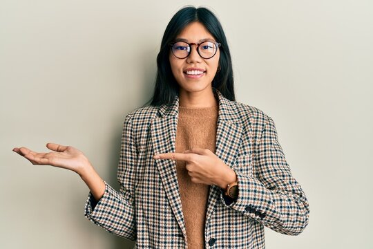 Young chinese woman wearing business style and glasses amazed and smiling to the camera while presenting with hand and pointing with finger.