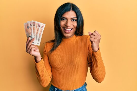 Young latin transsexual transgender woman holding 10 united kingdom pounds banknotes screaming proud, celebrating victory and success very excited with raised arm