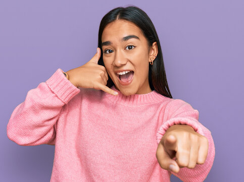 Young asian woman wearing casual winter sweater smiling doing talking on the telephone gesture and pointing to you. call me.