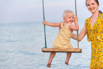 Young mother rolls the child on swing at the sea. Mom and child at the sea look at the camera and smile