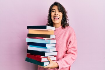 Young hispanic woman holding a pile of books smiling and laughing hard out loud because funny crazy joke.