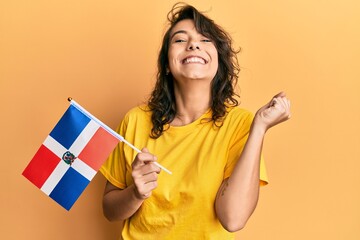 Young hispanic woman holding dominican republic flag screaming proud, celebrating victory and success very excited with raised arm
